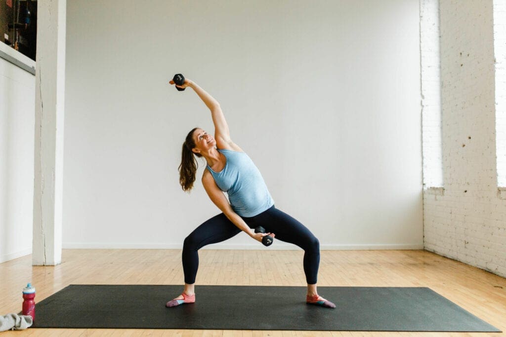 pregnant woman performing a pliè squat and overhead dumbbell reach in a barre class workout at home