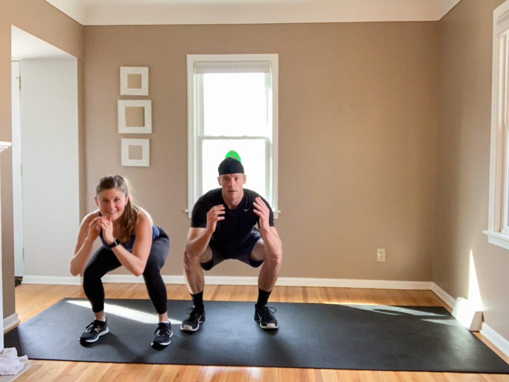 one woman and one man holding a bodyweight squat as part of a no equipment HIIT workout at home