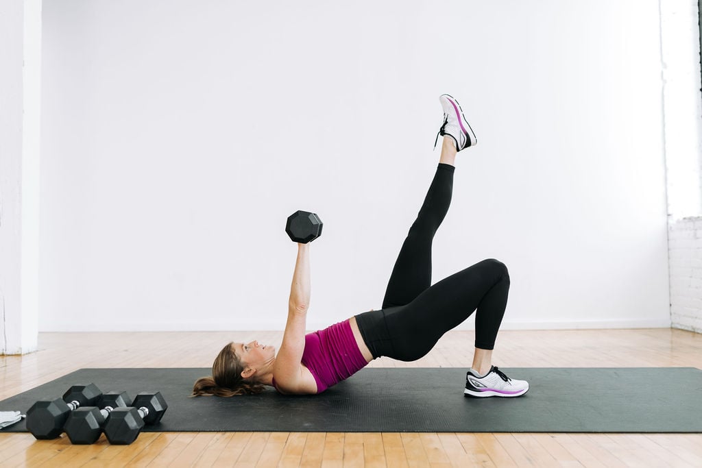 Woman performing a single leg glute bridge and chest press as part of a Legs and Chest Workout for Women At Home using weights