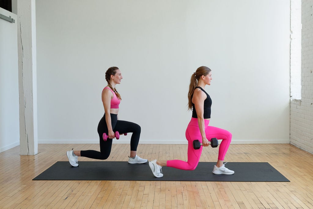 two women performing a reverse lunge with dumbbells in a conditioning workout at home