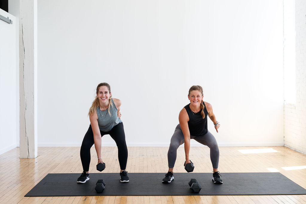 Two women performing a Dumbbell Squat, holding the dumbbell in their left hand. Postpartum Exercises to Build Strength At Home