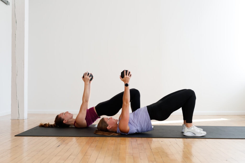 two women performing a glute bridge hold with dumbbell chest press in a first trimester workout
