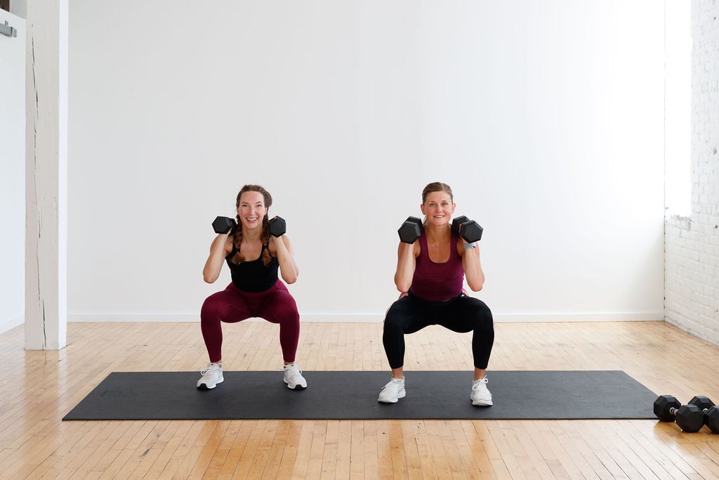 two women performing a squat hold with dumbbells front racked in a leg workout at home