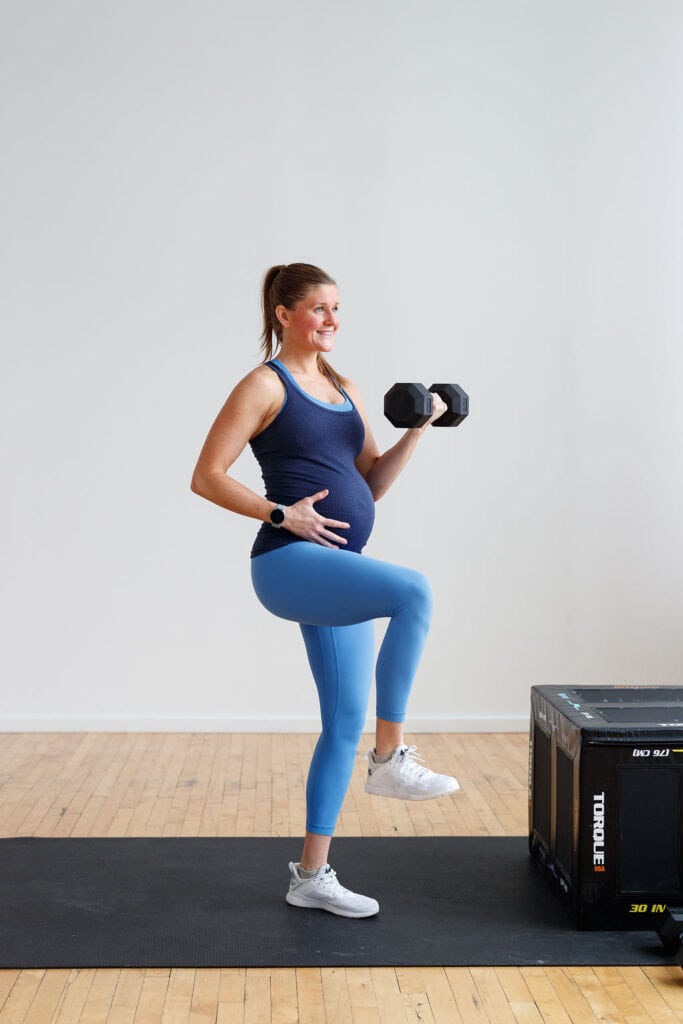 Pregnant women in light blue leggings and navy tank top doing a single leg bicep curl.