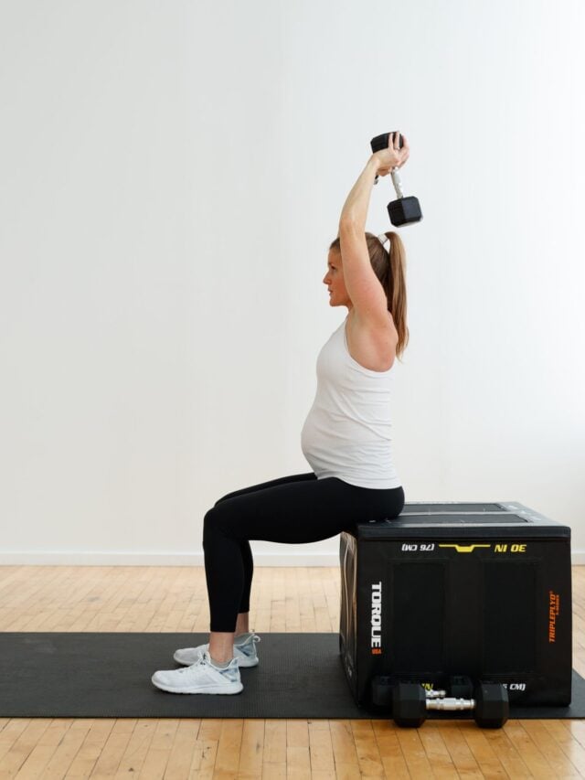 Pregnant woman sitting on a plyometric box performing a modified overhead tricep extension with a dumbbell.