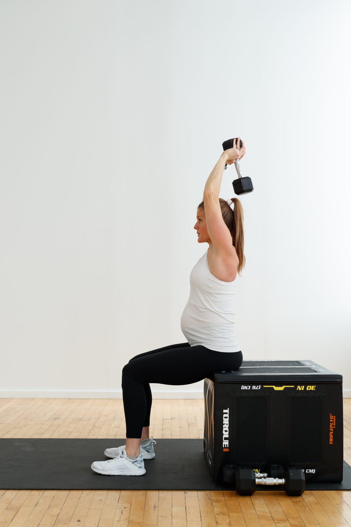 pregnant woman wearing white tank top and black maternity leggings performing a seated overhead tricep extension