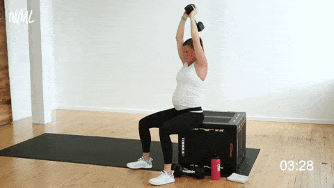 pregnant woman seated on a bench performing overhead tricep extensions with one dumbbell