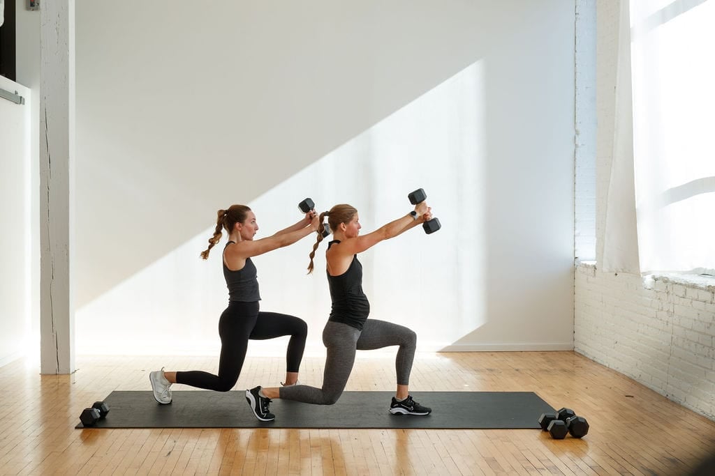35-Minute Leg and Shoulder Workout. Two women holding dumbbells performing a lunge and 45-degree dumbbell press.