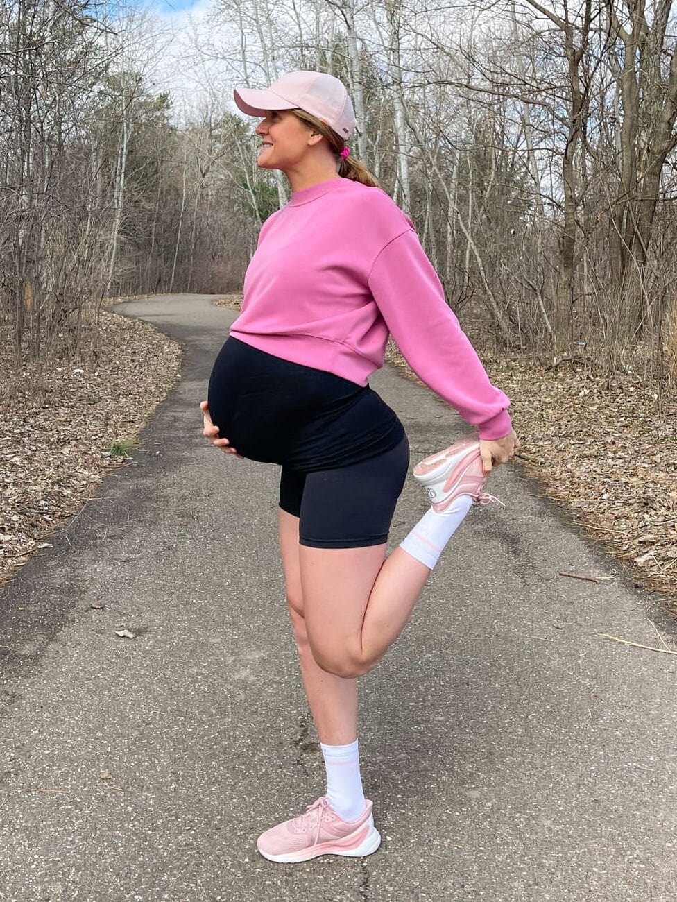 pregnant woman in pink sweatshirt wearing running shoes on a trail in the woods stretching 