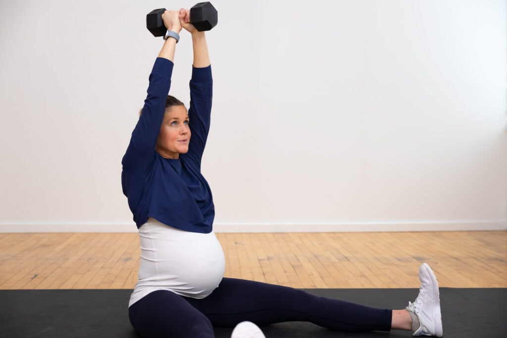 pregnant woman sitting on mat holding dumbbell overhead as part of prenatal core workout