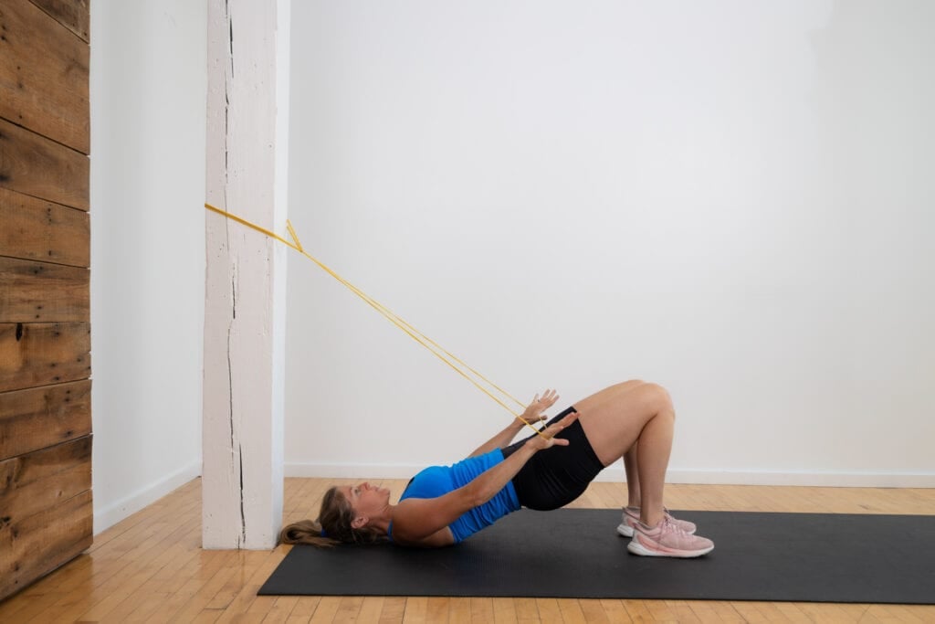 Postpartum woman performing a glute bridge with a band pull down as part of diastasis recti workout