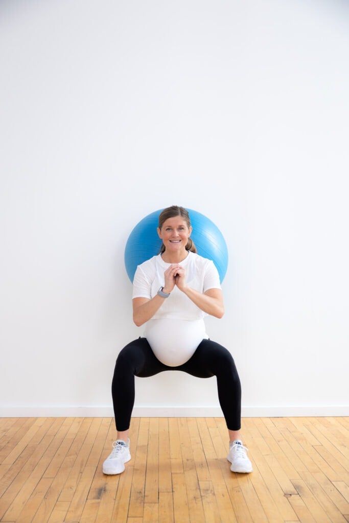 Pregnant woman performing a modified squat using an exercise and stability ball. The exercise and stability ball is a piece of top exercise equipment to provide modifications for a pregnancy fitness journey. 