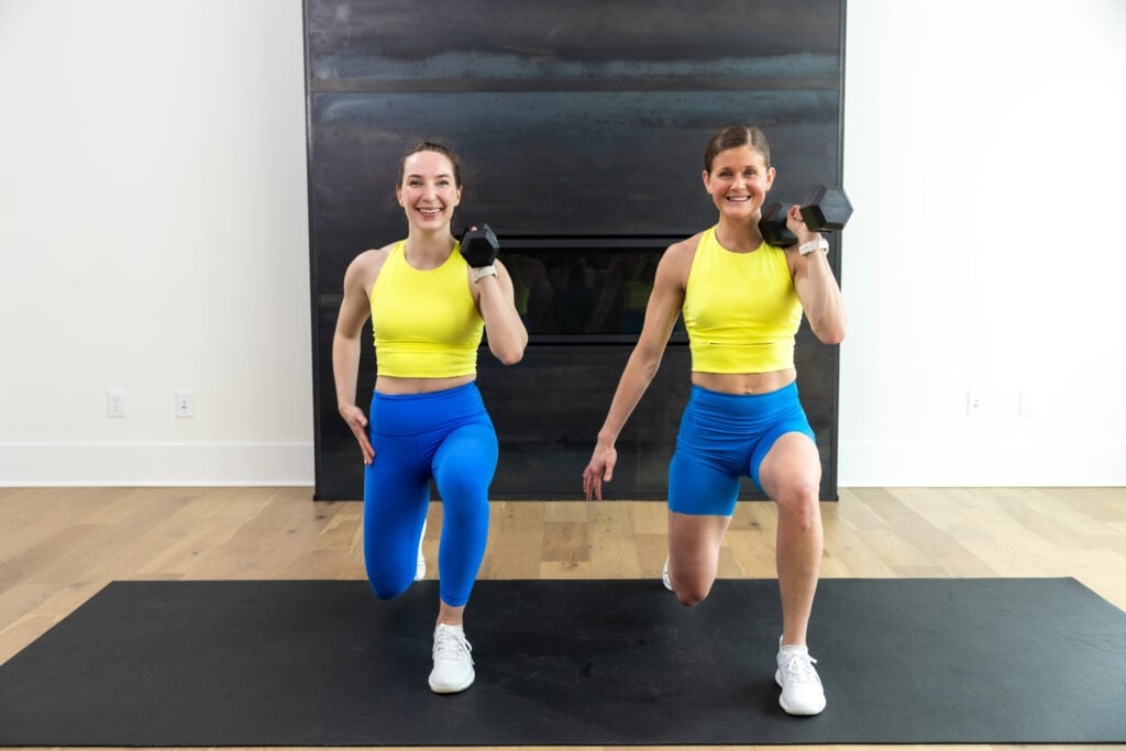 two women performing a reverse lunge and dumbbell hold, holding a single dumbbell in a front racked position at shoulder height in a full body push workout