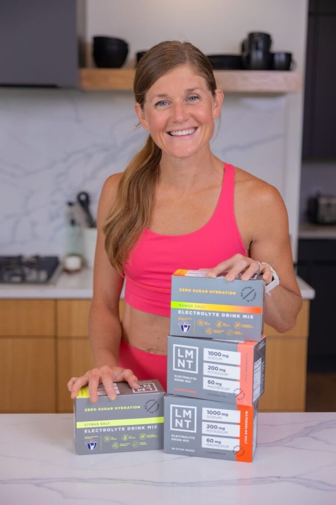woman standing in her kitchen next to several boxes of LMNT electrolyte drink mix | muscle recovery supplements