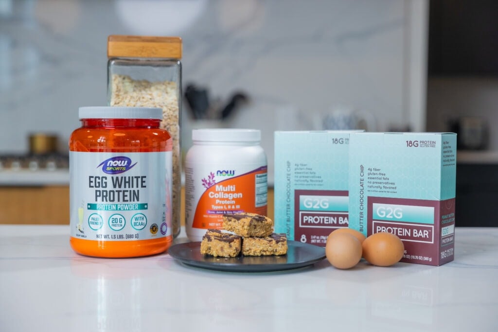 Five high protein snacks in a white kitchen, including egg white protein powder, eggs, collagen protein, oatmeal, peanut butter chocolate chip and mini peanut butter protein bars. 