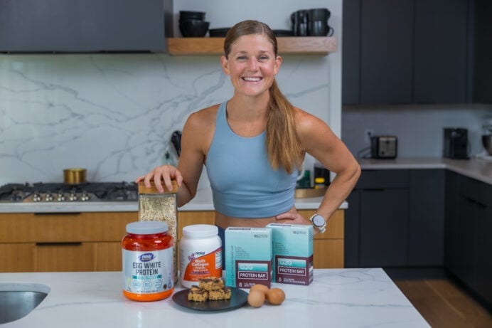 Women in kitchen with high protein snacks including protein powder, oatmeal, homemade peanut butter bars, eggs and protein bars.