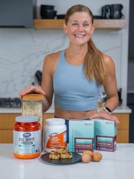 Woman in kitchen with high protein snacks including protein powder, oatmeal, homemade peanut butter bars, eggs and protein bars.