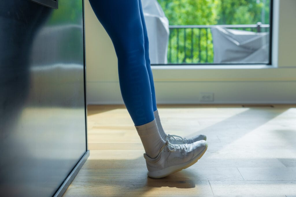 Woman performing tibialis raises to improve ankle mobility. Woman standing against a wall and pulling her toes toward her face as part of an ankle strengthening workout routine.