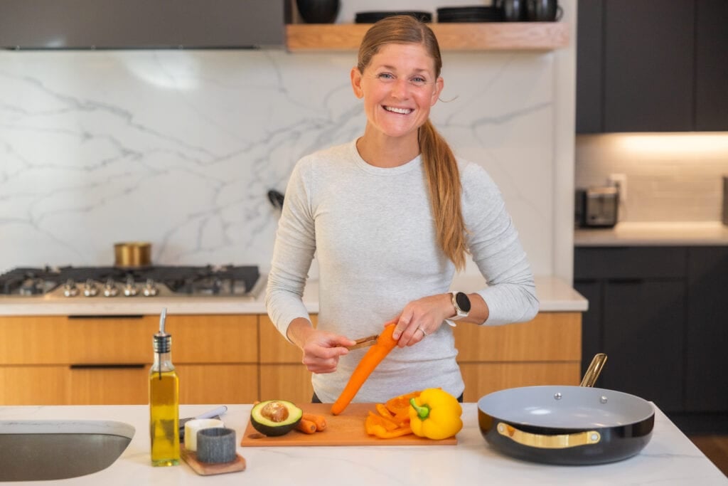 Women meal prepping in her kitchen; peeling a carrot on a cutting board with caraway fry pan next to her. 