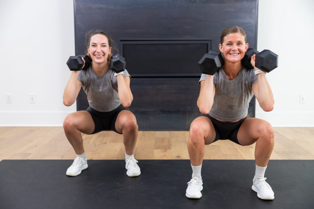 two women performing a squat with dumbbells front racked in a squat workout