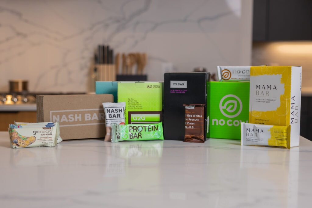 Boxes of protein bars lined up in a kitchen with brands including mama bars, gomarco bars, rxbars, g2g bars, nash bars and no cow protein bars.