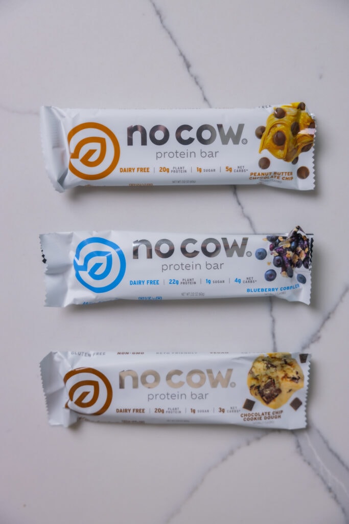 Three no cow protein bars with the flavors peanut butter chocolate chip, blueberry coboler, and chocolate chip cookie dough. 