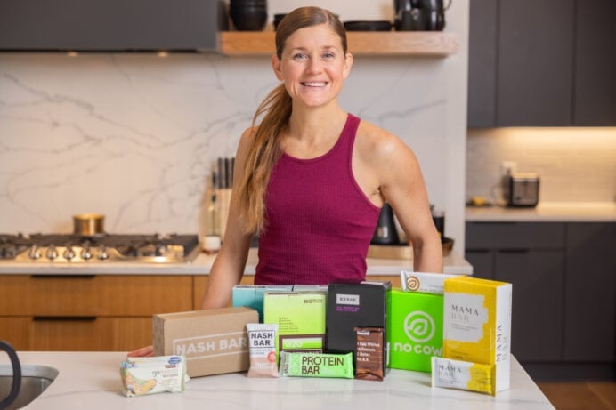 Women in her kitchen with brands of protein bars; no cow, g2g bars, rx bars, gomarco bars, mama bars and nash bars.