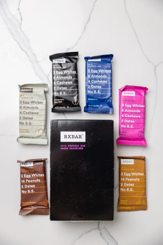 Box of RxBars protein bars and individual bars with flavors peanut butter chocolate chip, coconut chocolate, chocolate sea salt, blueberry, mixed berry and peanut butter. 
