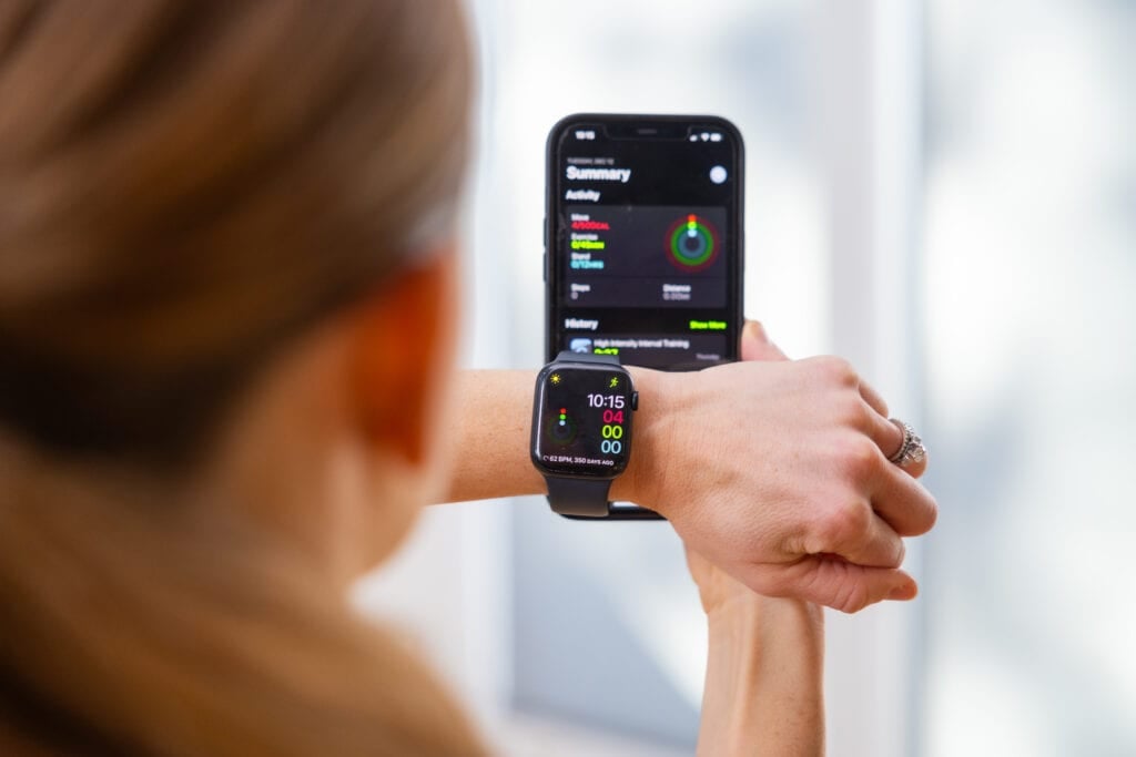Women wearing a black apple watch on her left wrist with the front screen on. Holding her phone with the apple watch app open in her right hand. 