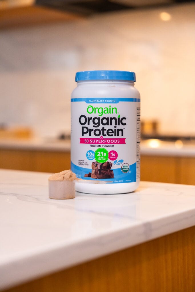 orgain organic chocolate protein protein powder on countertop with a scoop of chocolate protein as part of best protein powder