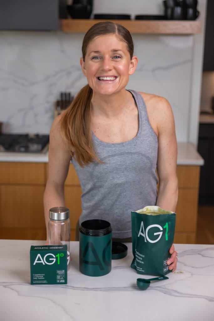 Lindsey in her kitchen with AG1 Greens including the greens powder, greens scoop, 5 travel pack of green powder, water bottle, and vitamin D with K3.