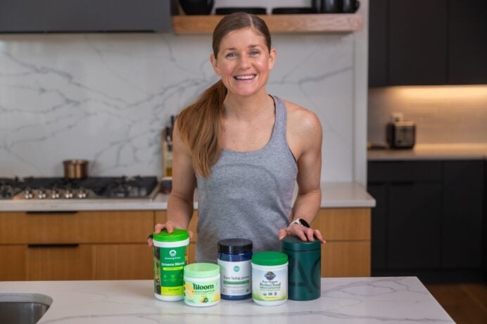 Women in her kitchen with the best greens powders including AG1, bloom greens, amazing grass greens, ora easy bring green, and garden of life greens.
