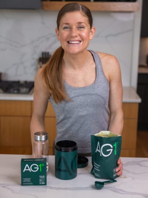Lindsey in her kitchen with AG1 Greens including the greens powder, greens scoop, 5 travel pack of green powder, water bottle, and vitamin D with K3.