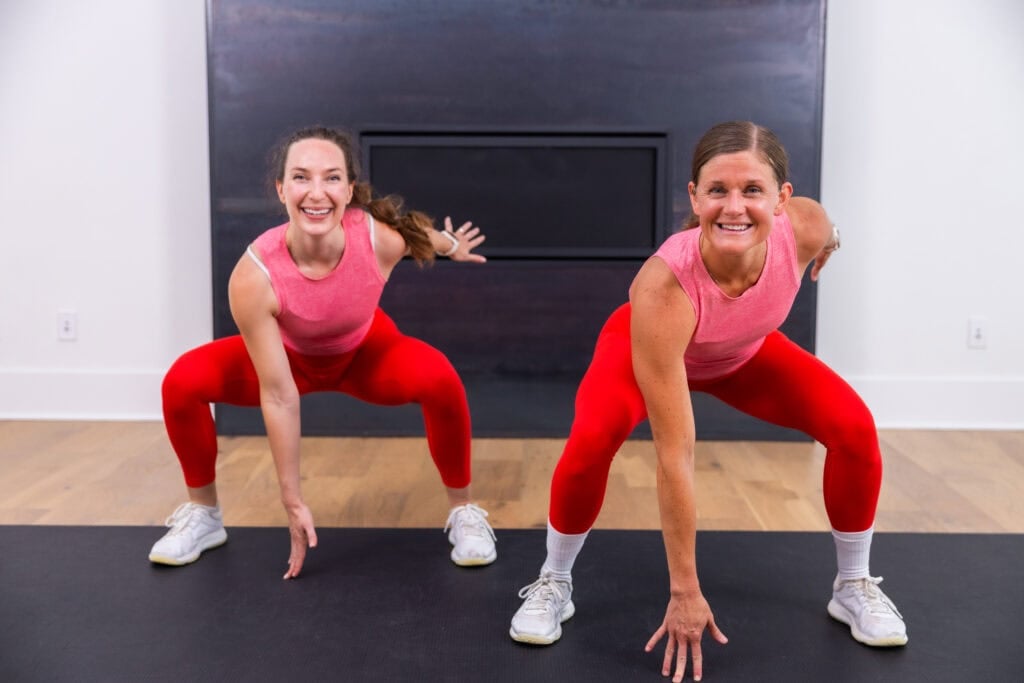 two women holding a low squat and tapping the ground at the bottom of a squat jack in a full body workout