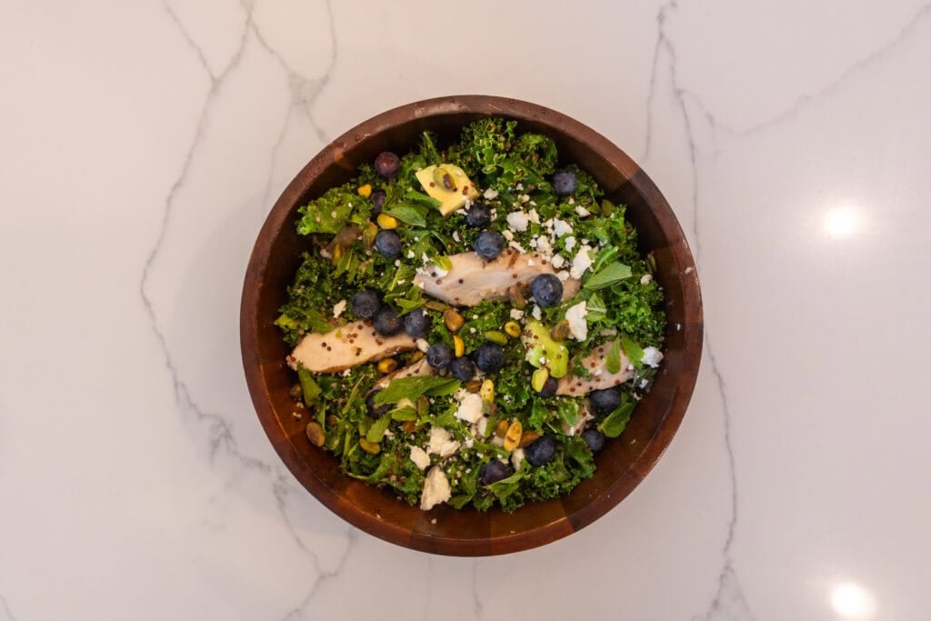 Salad recipe with kale, quinoa, blueberry, chicken, pumpkin seeds, feta cheese, avocado in a salad bowl.