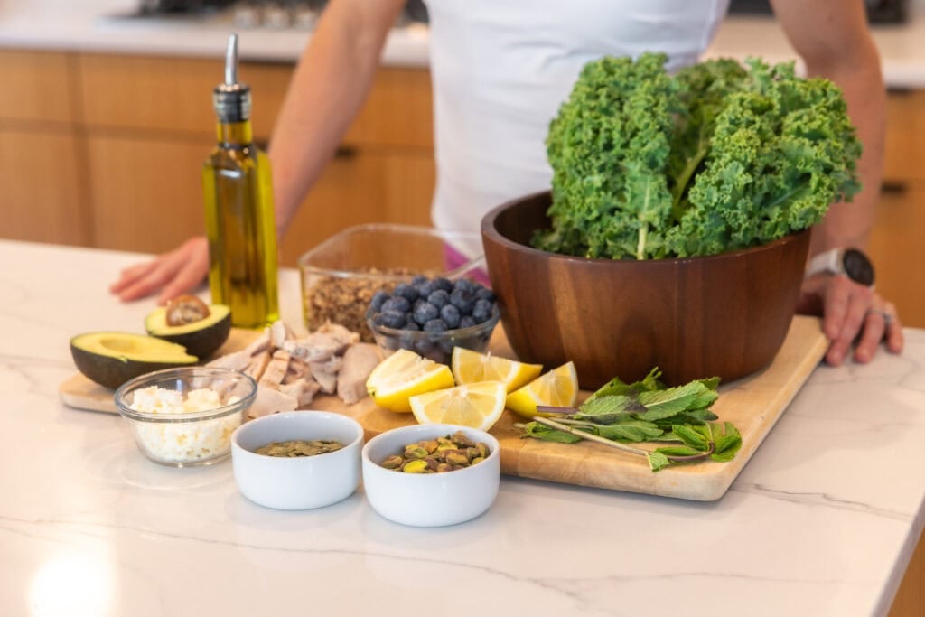 Salad ingredients including fresh kale, pumpkin seeds, feta cheese, mint, lemon juice, blueberry, chicken, avocado, quinoa and olive oil.