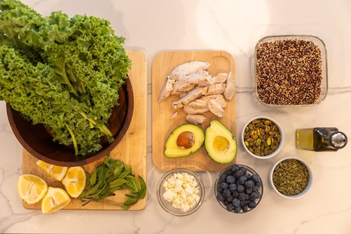 Salad ingredients including fresh kale, pumpkin seeds, feta cheese, mint, lemon juice, blueberry, chicken, avocado, quinoa and olive oil.