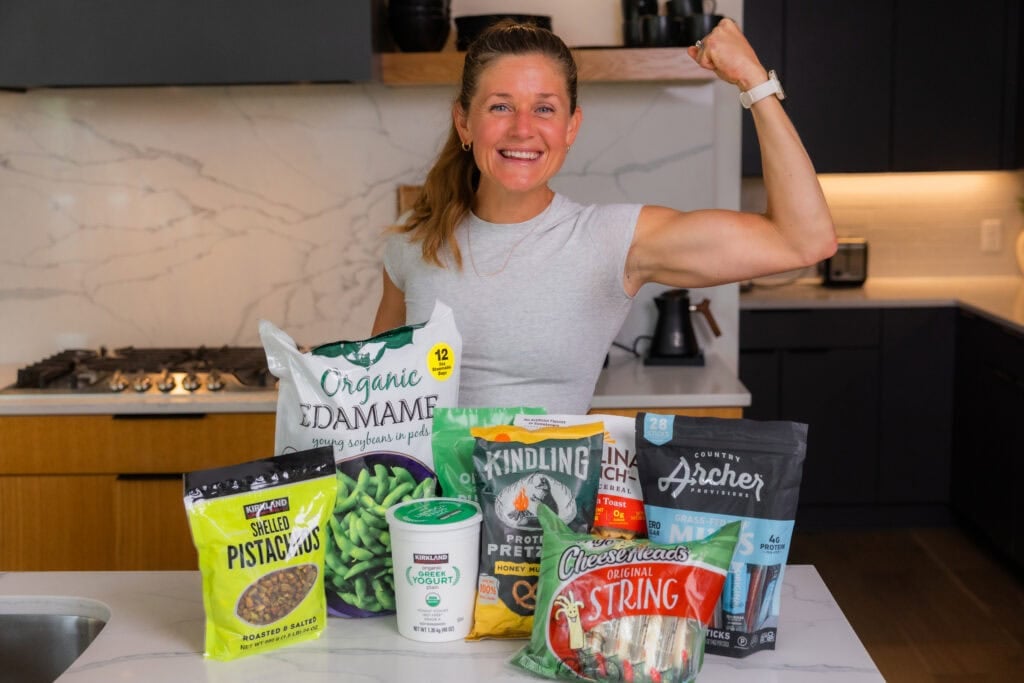 Women flexing with High protein costco snacks including refrigerated and Frozen Snacks, and Shelf-Stable and Pantry Snacks. 