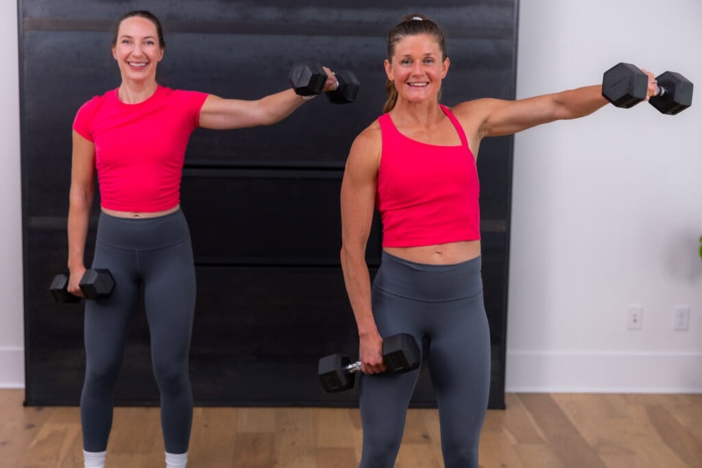 two women doing standing dumbbell lateral raises, one arm lifted to shoulder height in a home gym.