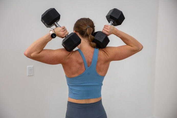 Woman holding two dumbbells in her hands over her shoulders in a blue workout top