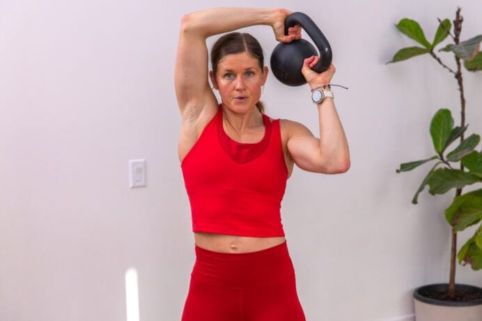A woman in a read workout outfit lifting a kettlebell over her head in a 20 minute beginner kettlebell workout.