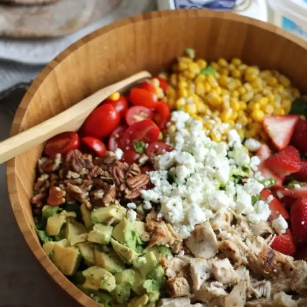 Summer Chicken Chopped Salad with greens, tomatoes, corn, avocado, strawberries, chicken, blue cheese in a wooden bowl.