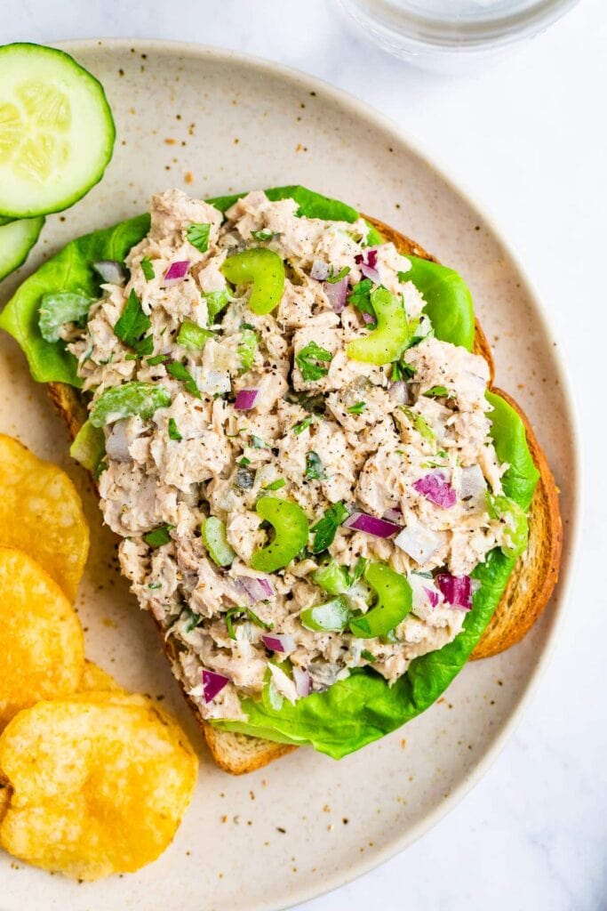 Tuna salad sandwich with green, tuna, celery, onion, bread, chips and cucumbers.