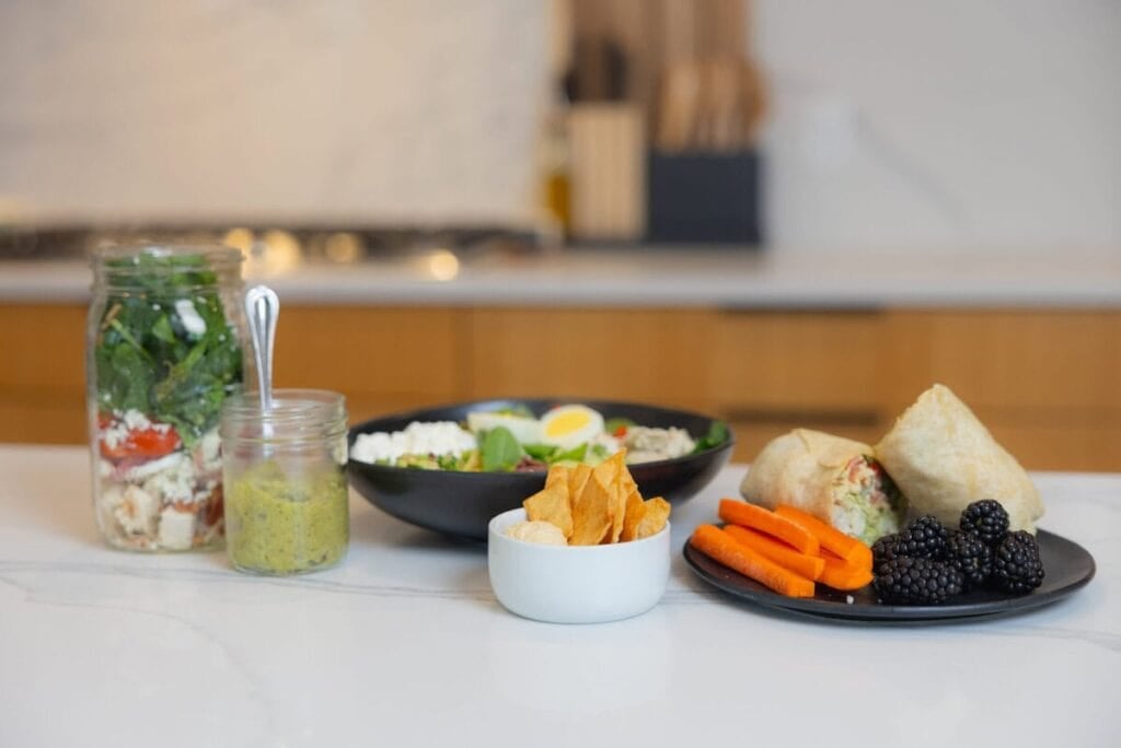 A spread of food with: a salad in a jar, a Cobb salad, a Caesar chicken wrap with carrots and blueberries, pita chips with hummus