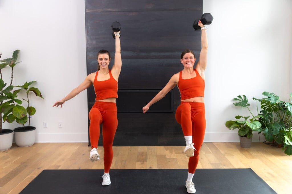 Two women doing a Knee Drive Shoulder Press with one dumbbell. 