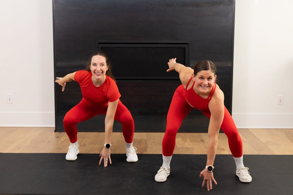 Two women in red workout clothes performing a wide squat with a rotational reach during an indoor workout.